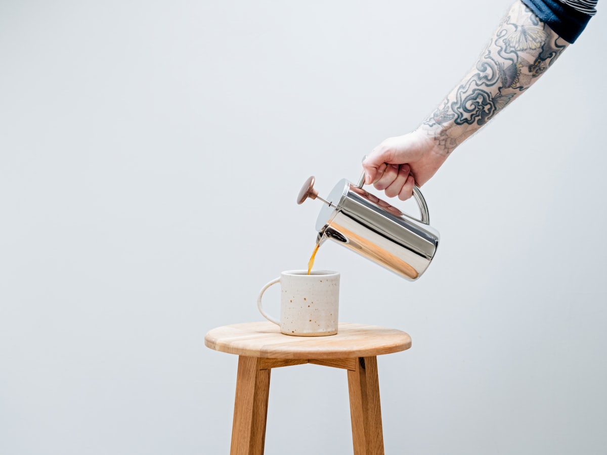 A hand pouring hot water over coffee grounds from a kettle into a mug on a wooden stool